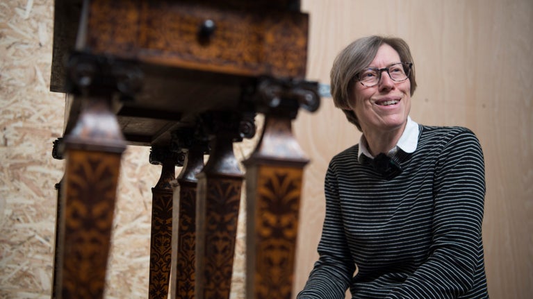Sophie Chessum dressed in a black jumper and kneeling besides a marquetry table.